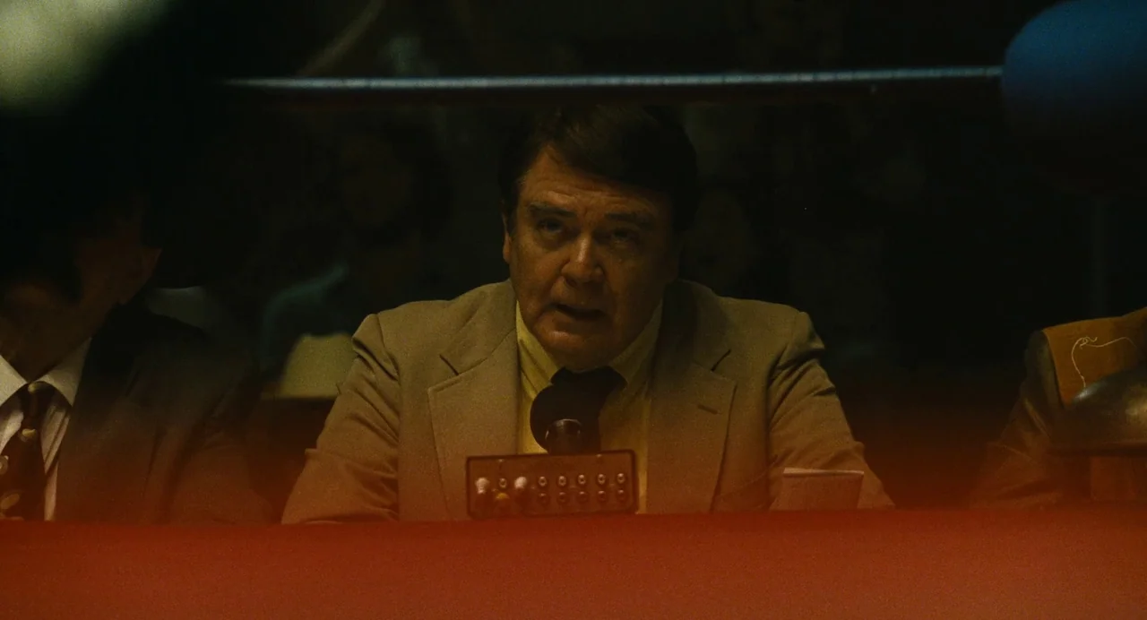 The Iron Claw (2023) movie still: medium — A man sits at a broadcast table featuring a control panel with knobs. He leans into a microphone while…