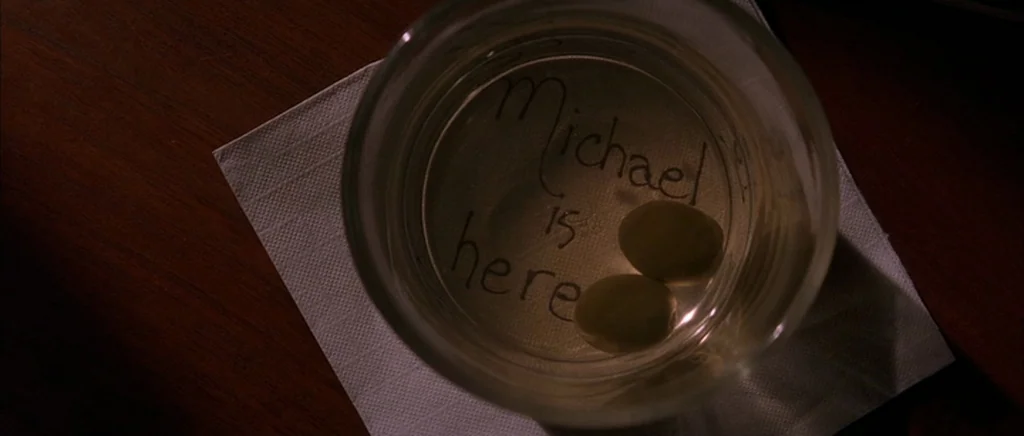 The Underneath (1995) movie still: closeup — A glass of liquid with two olives rests on a white paper napkin. Handwritten text on the bottom of the…