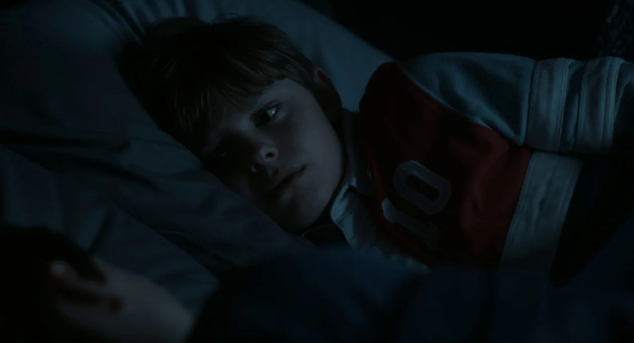 Goodnight Mommy (2022) movie still: closeup — A dark bedroom at night features a child lying on a pillow. The child watches another person while…
