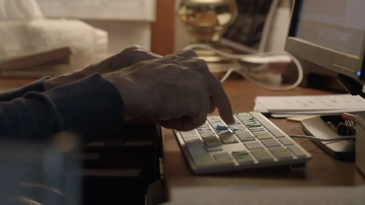 Breslin and Hamill: Deadline Artists (2018) movie still: closeup — A wooden desk holds a computer keyboard and a monitor in a bright room. The hands…