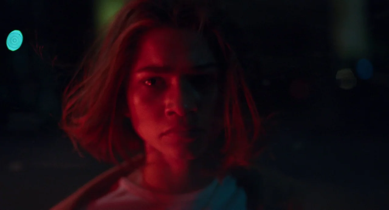 Challengers (2024) movie still: closeup — A dark city street at night with out-of-focus street lights in the background. A young woman with…