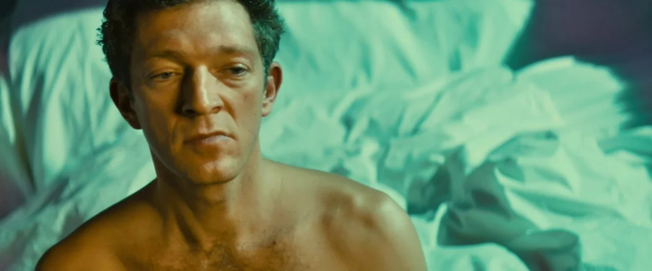 Trance (2013) movie still: closeup — A man sits shirtless on rumpled white bed sheets inside a dark room. The subject looks forward with a neutral…