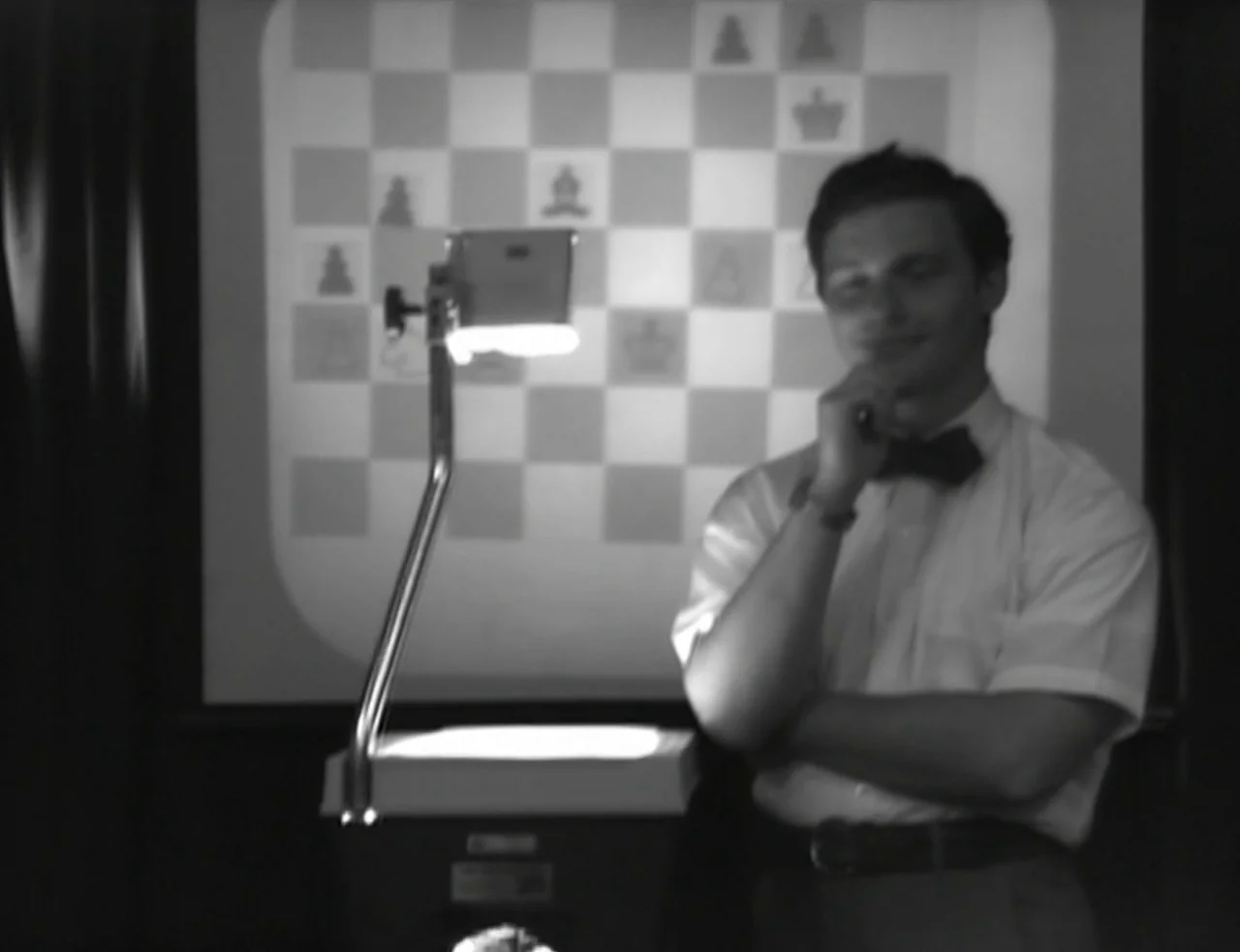 Computer Chess (2013) movie still: medium — A darkened room features a projection screen displaying a large translucent chess board image. A man…