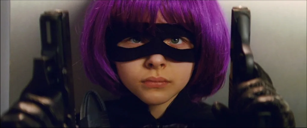 Kick Ass (2010) movie still: closeup — A close up shows a teenage girl with purple hair holding two handguns forward. The subject stares directly at…