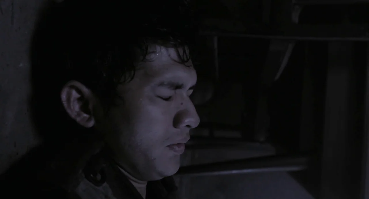 The Raid (2012) movie still: closeup — A person rests their head against a dark, featureless wall in low lighting. The man has eyes closed while…