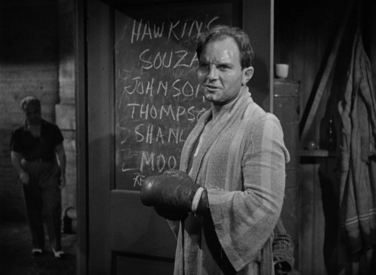 The Set-Up (1949) movie still: medium — A dimly lit locker room features a doorway with a list of names written. A muscular man in a robe and boxing…