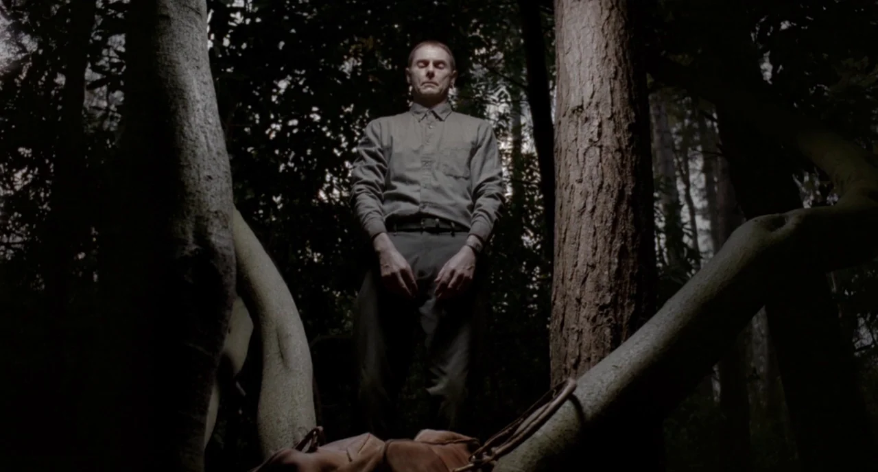 Possum (2018) movie still: fullbody — A man stands between two trees in a dense woodland area with dark foliage. The subject stands still with his…