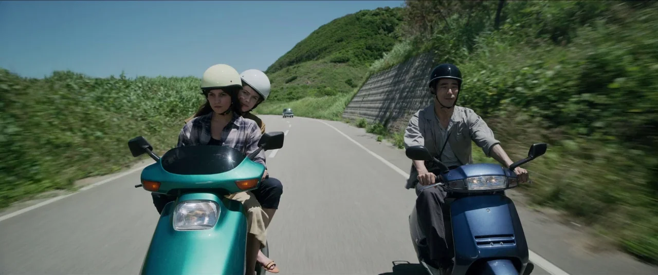 Earthquake Bird movie still: medium — An asphalt road winds through a sunny, green-covered coastal hill landscape during the day. Three people ride…