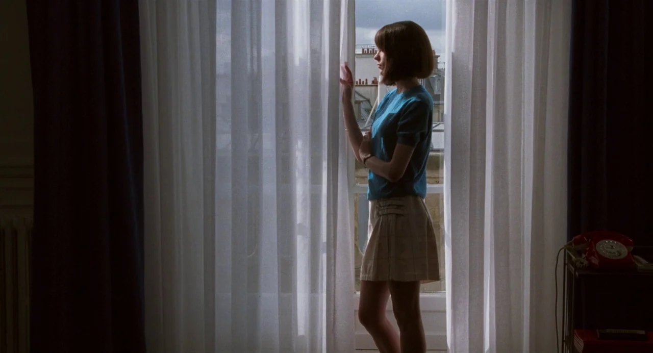 Godard Mon Amour (2015) movie still: medium — A woman in a blue sweater stands before a large window draped in sheers. Outside, a cityscape with…
