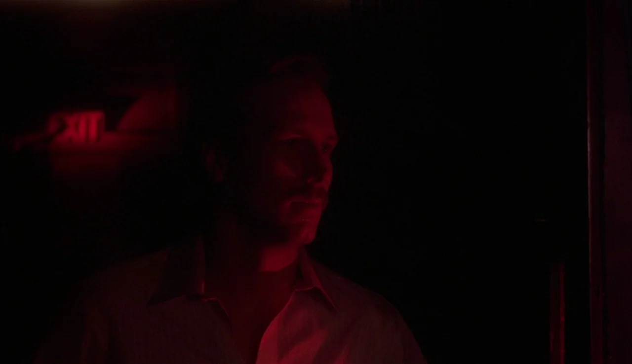 Body Heat (1981) movie still: medium — A man stands in a near-total darkness illuminated by a red exit sign. He looks toward the side while…