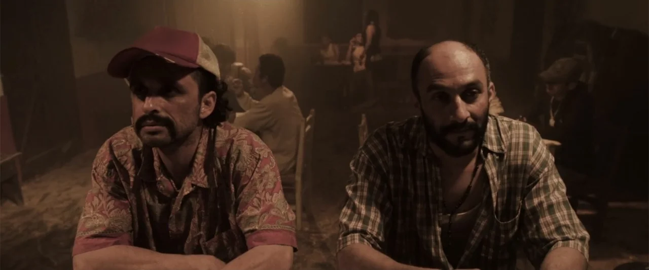 At Dawn (2013) movie still: medium — A dimly lit indoor bar with rustic wooden furniture and patrons in the background. Two men sit at a table in…