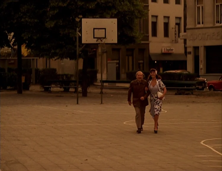 The Arrival of Joachim Stiller (1976) movie still: fullbody — A paved area with a basketball hoop structure stands in front of city buildings. An…