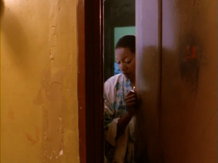 Tenement (1985) movie still: medium — A dimly lit narrow hallway with yellow plaster walls showing signs of age. A woman in a robe stands behind an…