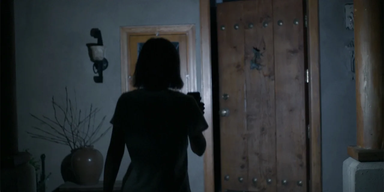 Demonic (2015) movie still: medium — A dimly lit interior hallway features a solid wooden door and a decorative vase. A person with shoulder-length…
