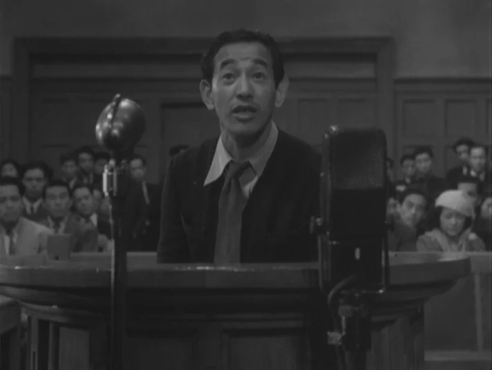 Scandal (1950) movie still: medium — A man speaks at a wooden witness stand in a crowded wood-paneled room. Multiple people sit in the background…