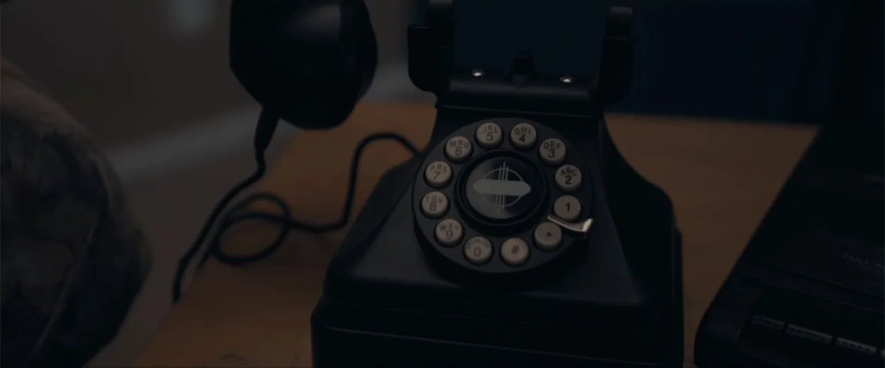 Anomaly (2006) movie still: closeup — A black vintage rotary telephone sits on a wooden surface in a dark room. The device occupies the frame center…