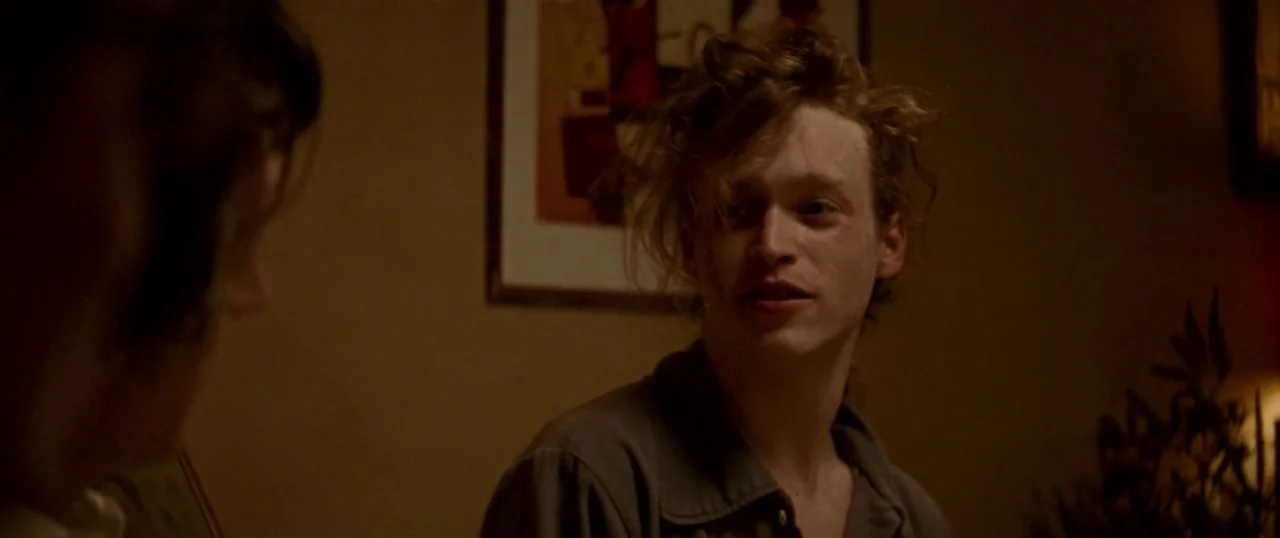 Low Down (2014) movie still: medium — A dimly lit interior room contains a framed abstract art piece on the wall. A young man with messy hair sits…