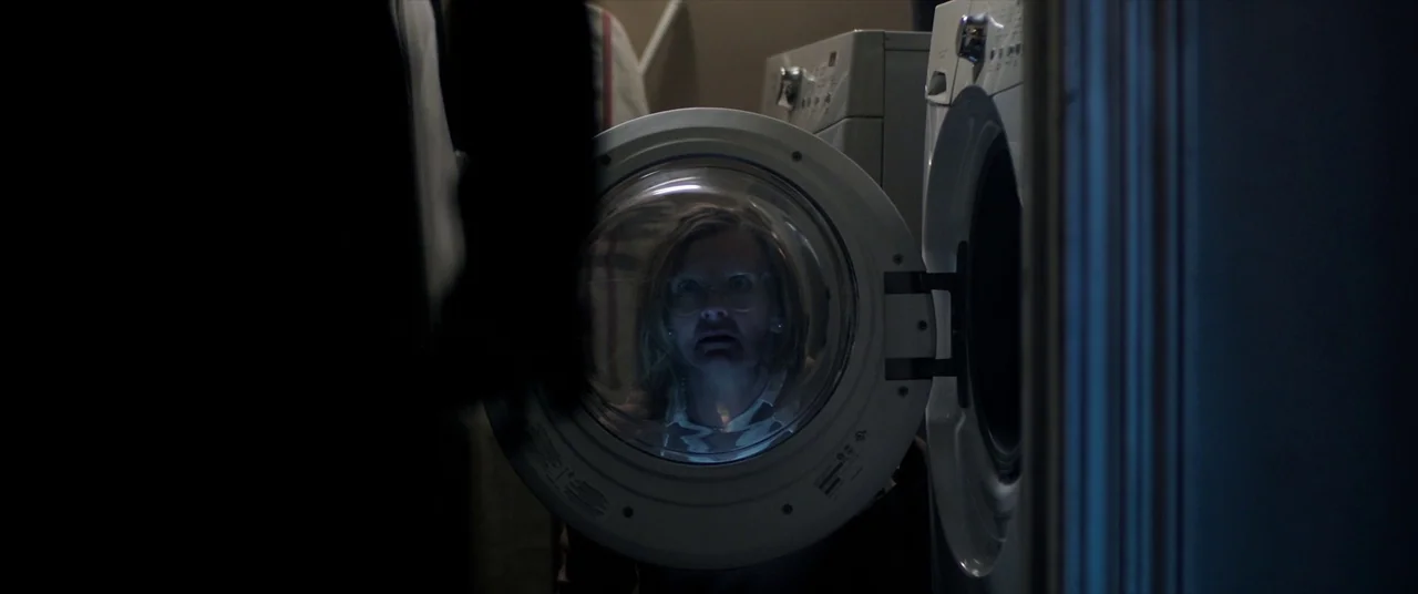 Malignant (2021) movie still: medium — A dimly lit laundry room features white appliances and a dark foreground figure. A woman peers through the…