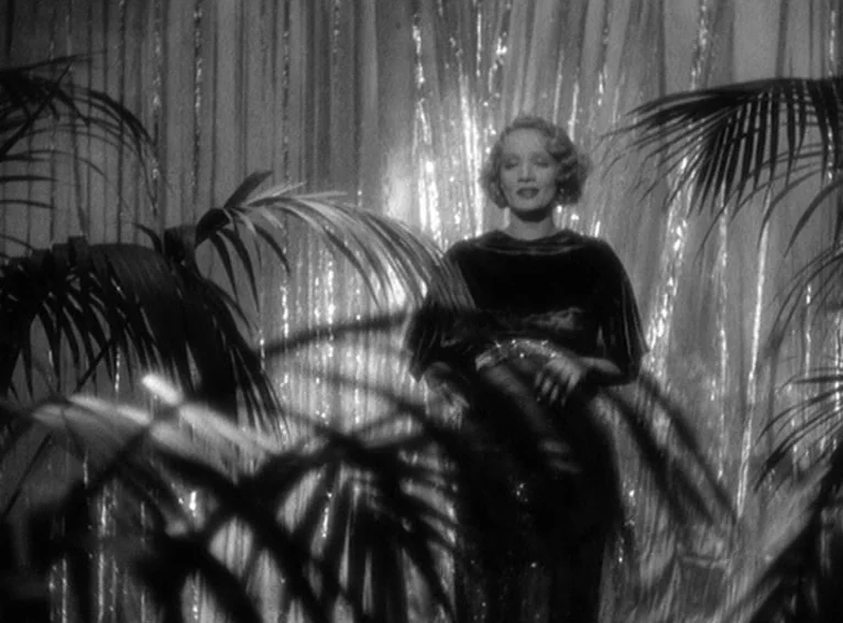 Blonde Venus (1932) movie still: medium — A woman stands on a stage in front of a backdrop of shimmering vertical metallic strips. Large palm leaves…