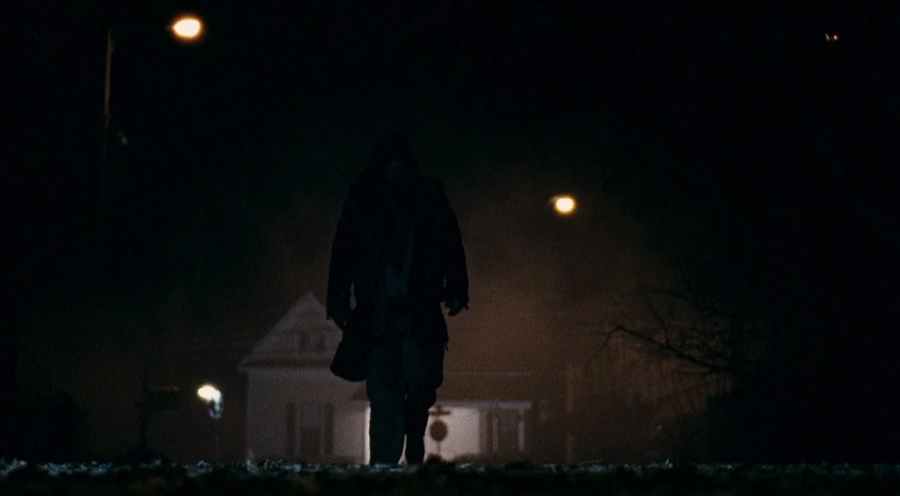 Halloween 2 (2009) movie still: fullbody — A dark residential street at night features a solitary figure walking toward the camera. A house is…