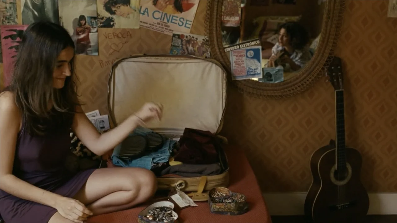 I'm Still Here (2024) movie still: medium — A bedroom features patterned brown wallpaper covered with posters and a round woven mirror. A woman in a…