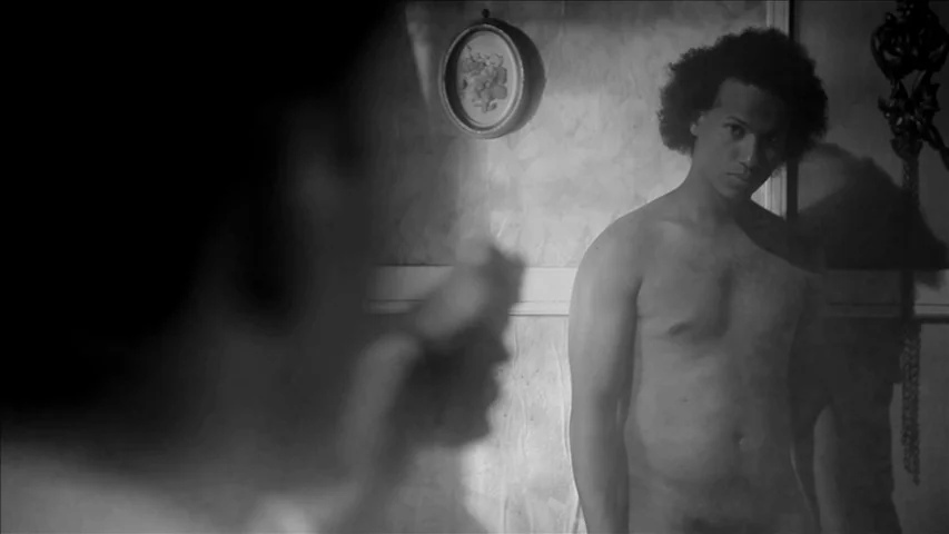 Keyhole (2011) movie still: medium — A dimly lit room features a patterned wall with an oval-framed picture. A shirtless man stands facing an…