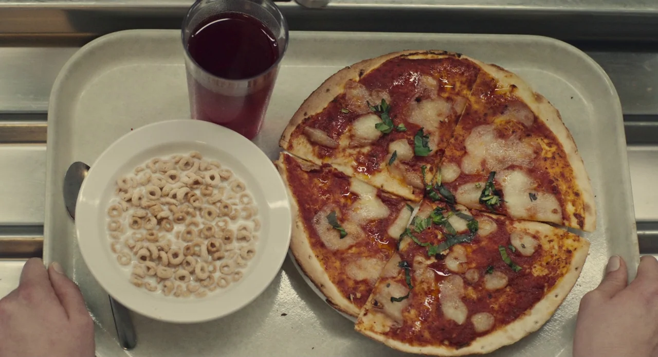 Mistress America (2015) movie still: closeup — A plastic cafeteria tray holds a small white bowl of cereal, a pizza, and a drink. A person's hands…