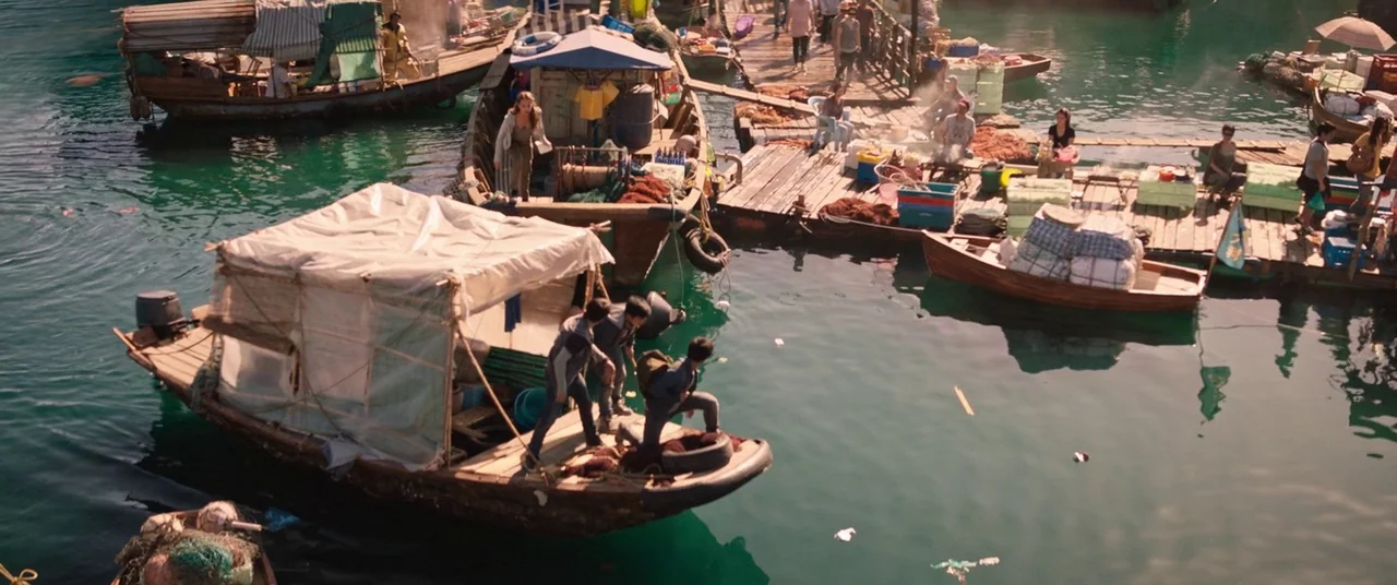 Tomb Raider (2018) movie still: wide — A busy harbor with wooden docks and numerous small fishing boats filled with nets. Three teenagers stand on…
