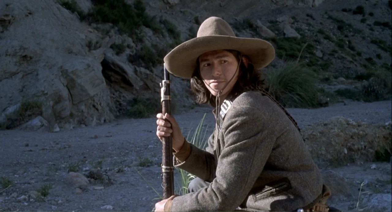 Chato's Land (1972) movie still: medium — A rocky canyon floor with a high limestone cliff face in the background. A young man in a wide-brimmed hat…
