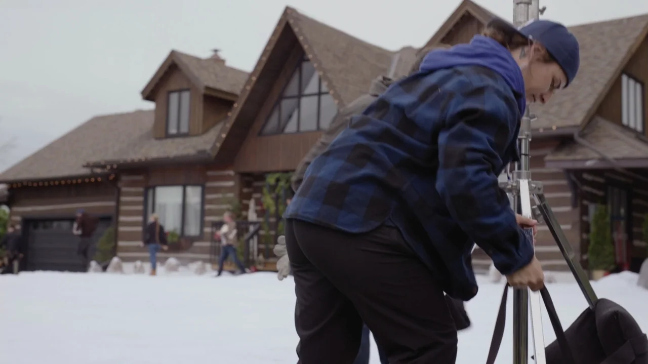 Designing Christmas (2022) movie still: medium — A snow-covered front yard stands before a large modern log cabin house. A person adjusts a metal…