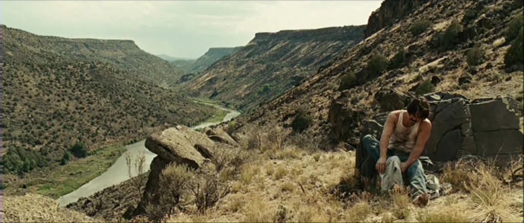 No Country For Old Men movie still: wide — A deep arid canyon stretches out with a river winding through the valley floor. A man sits on a large…