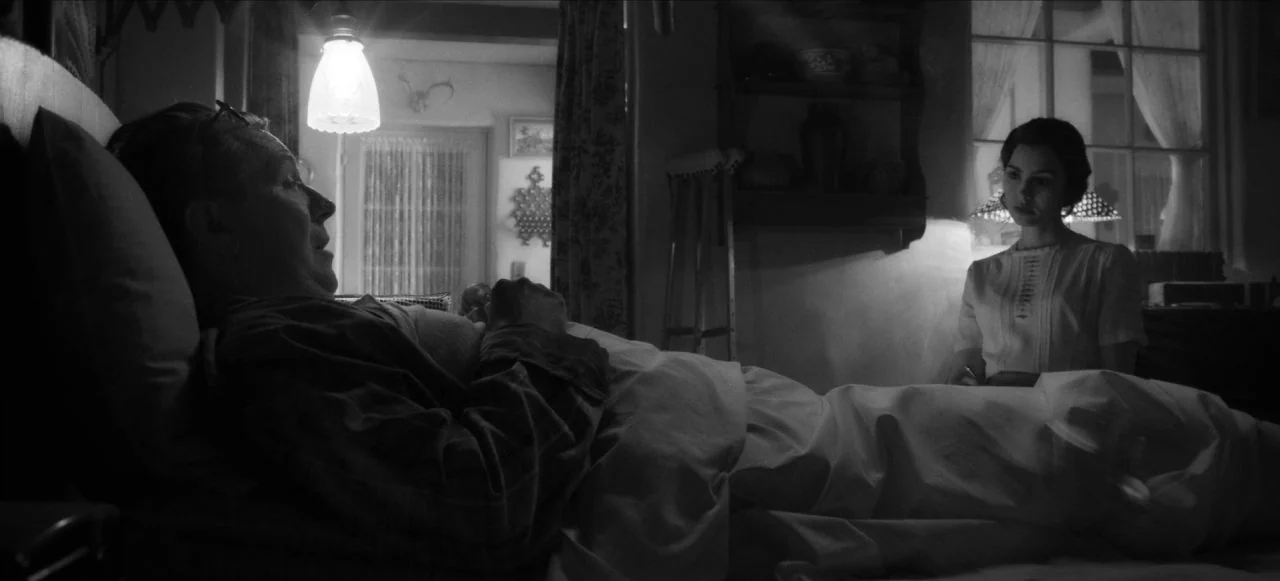 Mank (2020) movie still: medium — A dimly lit room contains an elderly person in bed and a young caregiver. The background features a window with…
