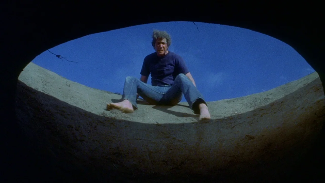 Phase IV (1974) movie still: fullbody — A man sits at the edge of a circular earthen hole looking down. The sky is bright blue and the ground is dry…