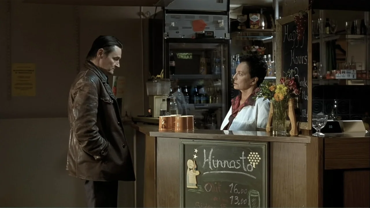 The Man Without a Past (2002) movie still: medium — A quiet bar interior features a wooden counter and a chalkboard menu display. A man in a brown…