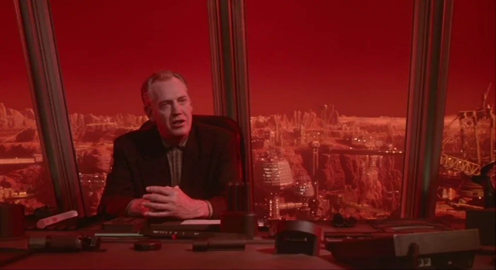 Total Recall (1990) movie still: medium — A futuristic high-rise office overlooking a red, rocky, industrialized alien landscape. A man sits at a…
