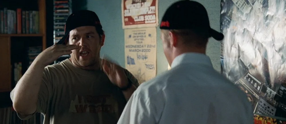 Shaun Of The Dead (2004) movie still: medium — A cluttered retail space with posters on a wall and shelves in the background. A man in a tan shirt…