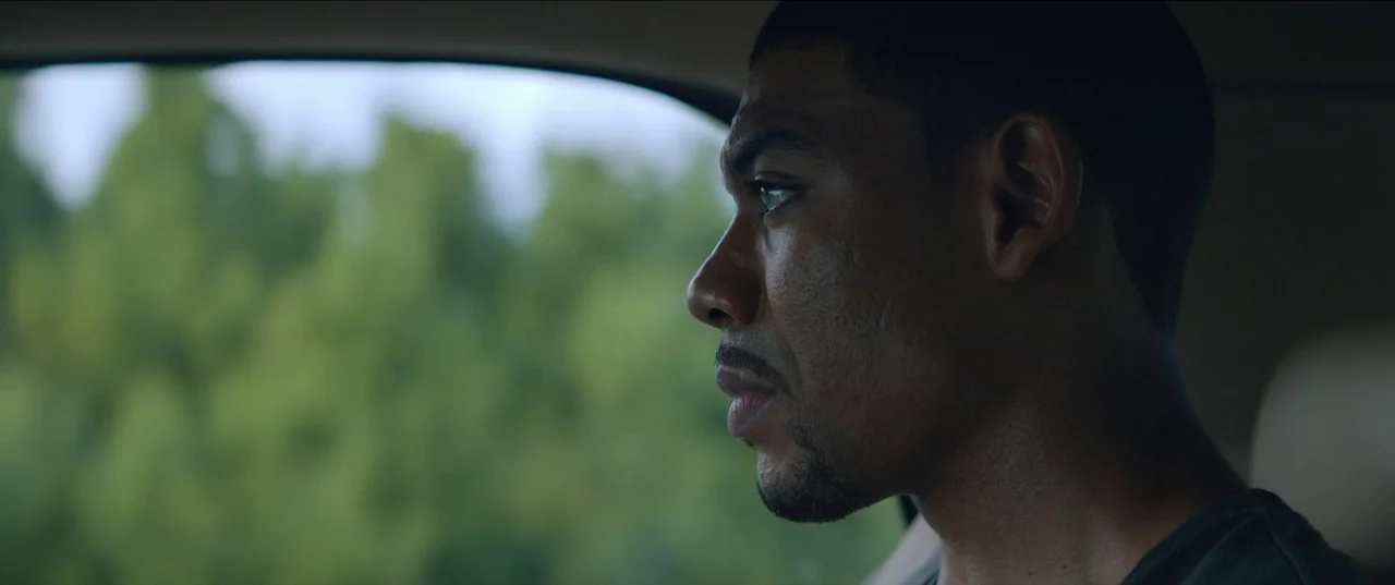Rebel Ridge (2024) movie still: closeup — A man in a dark shirt sits in the passenger seat of a car. He looks through the window at blurred green…
