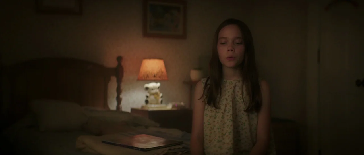 Pete’s Dragon (2016) movie still: medium — A dimly lit room with floral wallpaper and a bed in the background. A young girl sits facing the camera…