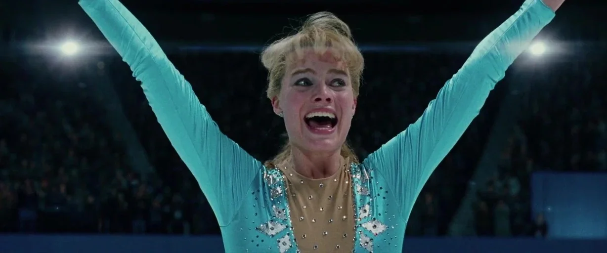 I, Tonya (2017) movie still: medium — A brightly lit indoor ice skating rink filled with a blurred spectator crowd. A woman in a teal skating outfit…