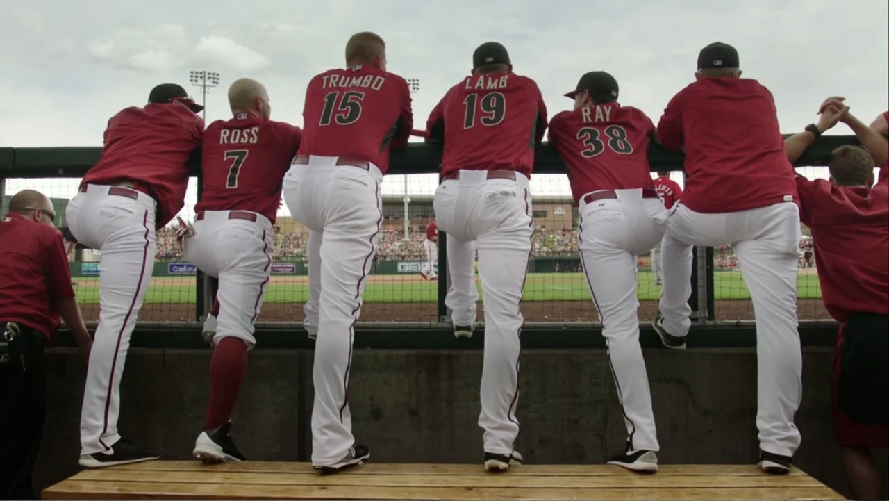 Ferrell Takes the Field (2015) movie still: fullbody — A baseball dugout bench sits in front of a chain-link fence at a stadium. Several…