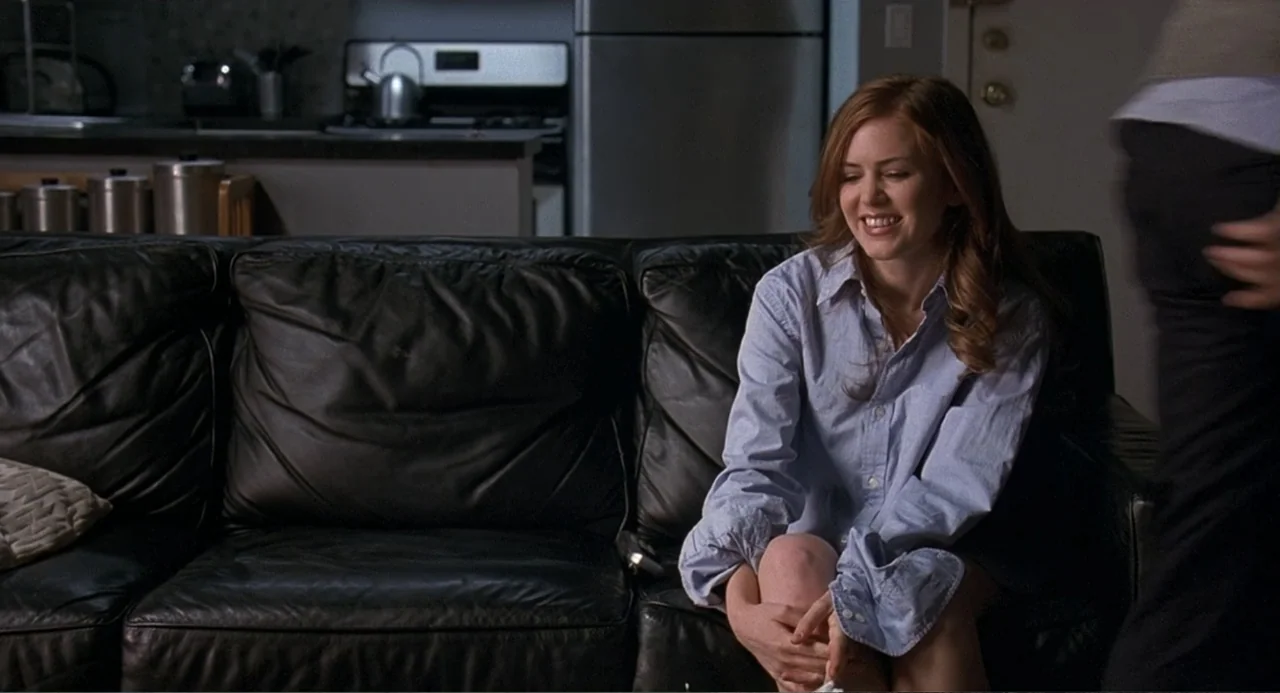 Wedding Daze (2006) movie still: medium — A modern living room with a black leather couch and kitchen in background. A young woman in a button-down…