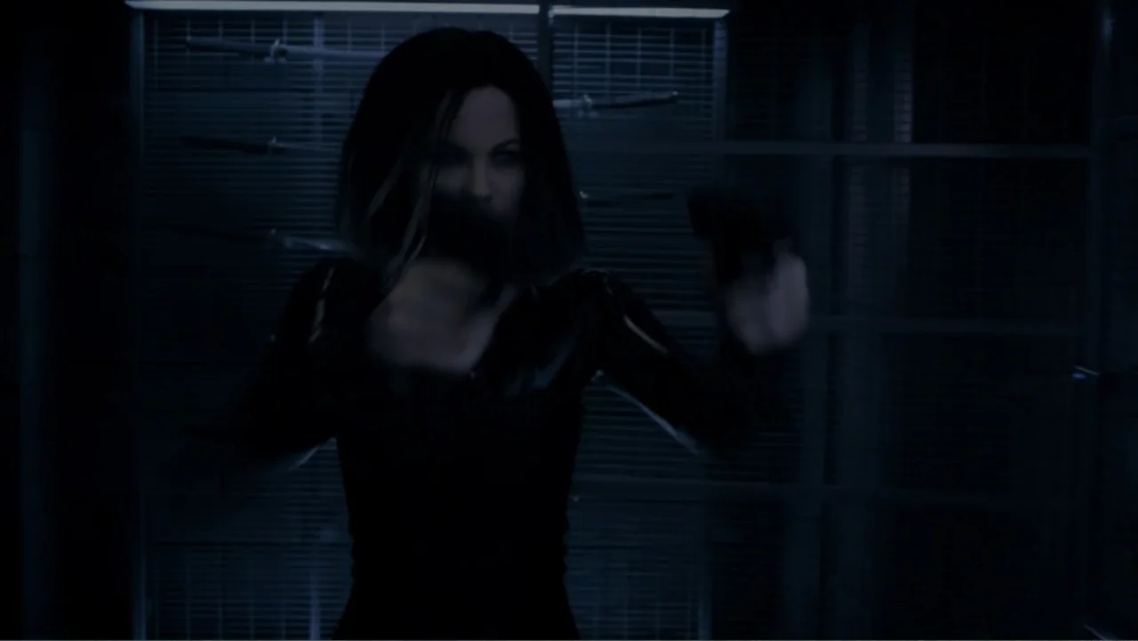 Underworld: Blood Wars (2016) movie still: medium — A dimly lit room features a metal grate backdrop with various weapons mounted behind. A woman in…