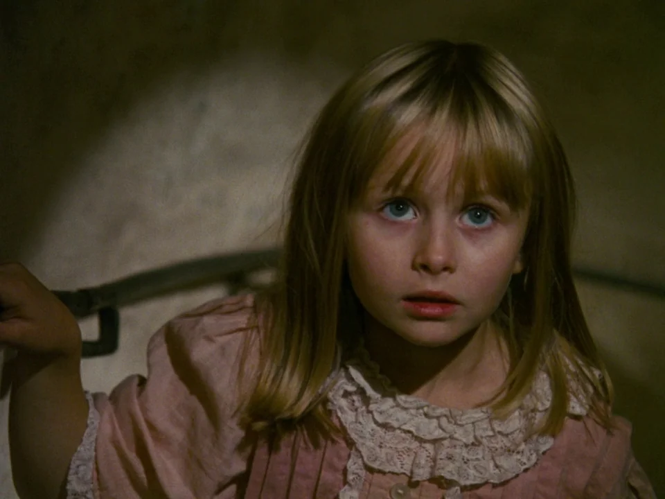 Alice (1988) movie still: closeup — A dimly lit interior staircase provides a dark, enclosed backdrop for the child. The young girl stares ahead…