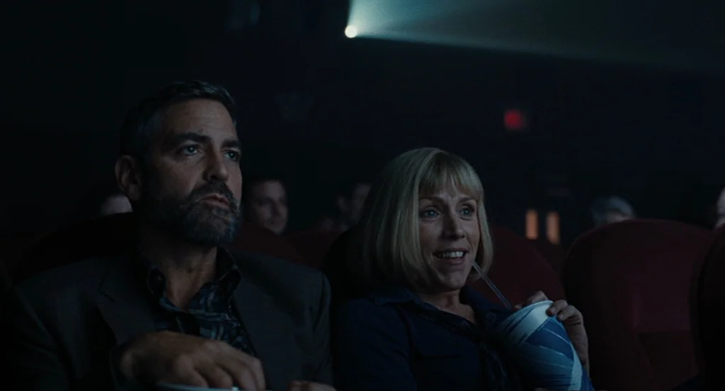 Burn After Reading (2008) movie still: medium — Two individuals sit in a dark movie theater with red velvet seating visible. A man looks ahead while…