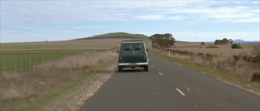 Roadgames (1981) movie still: wide — A dark green van travels down a desolate two-lane asphalt road through open fields. Fences line the roadside…