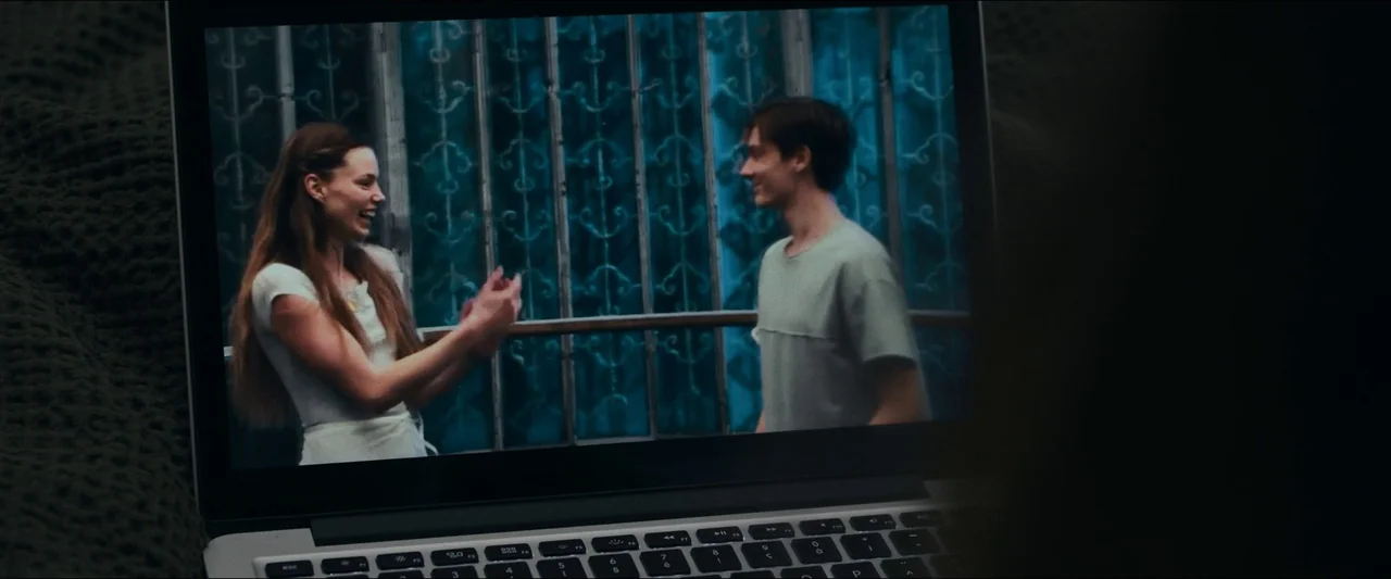 Birds of Paradise (2021) movie still: medium — A silver laptop sits on a dark surface, displaying a video of two people. The video shows a woman and…