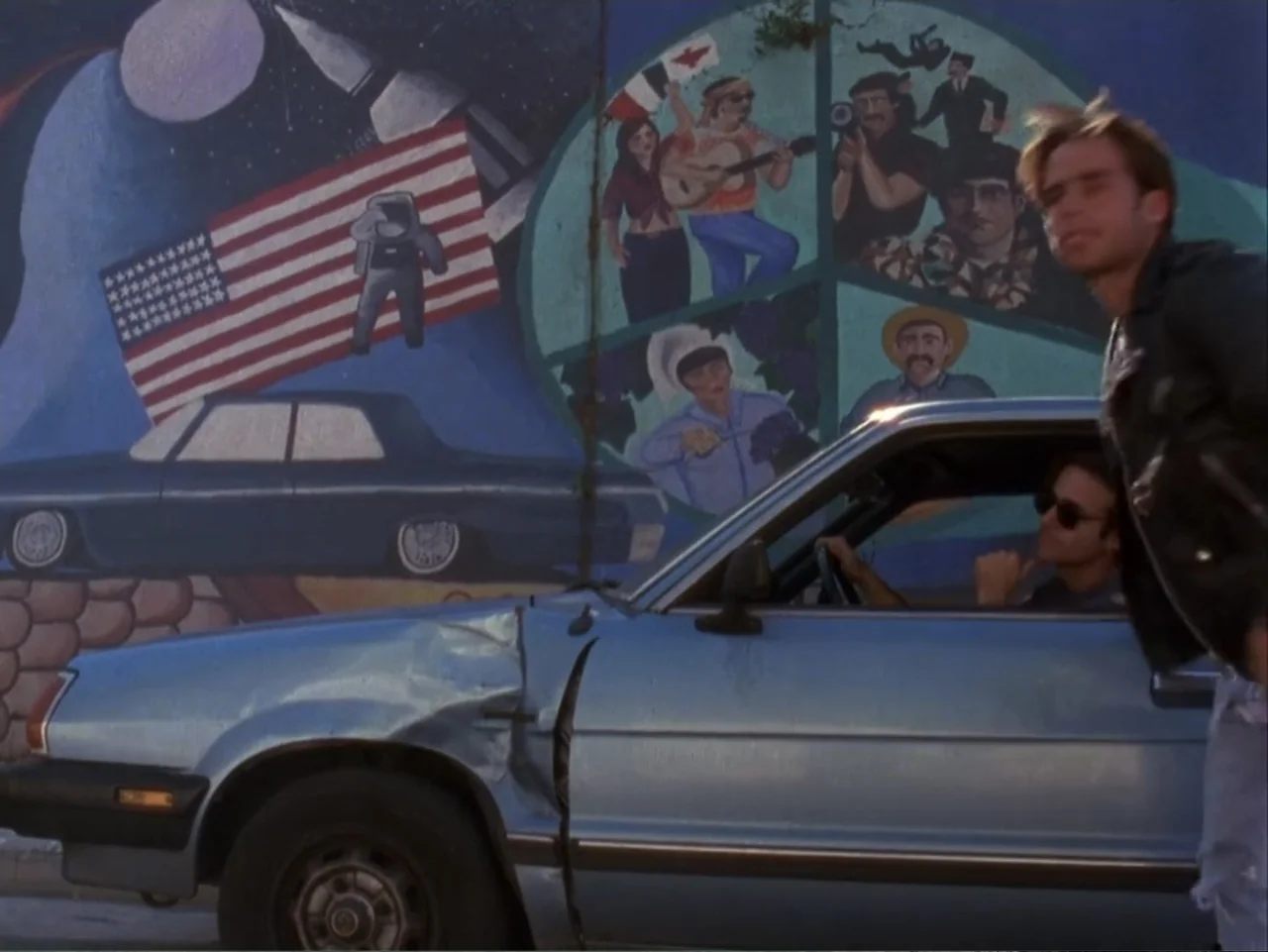 The Living End (1992) movie still: medium — A light blue damaged car is parked against a wall featuring a large colorful mural. Two young men are…