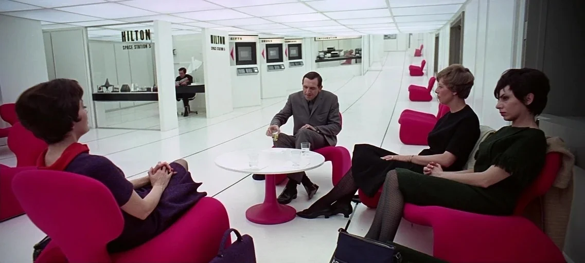 2001: A Space Odyssey (1968) movie still: medium — A stark white, sterile space station lounge features bright grid-patterned ceiling lighting. Four…