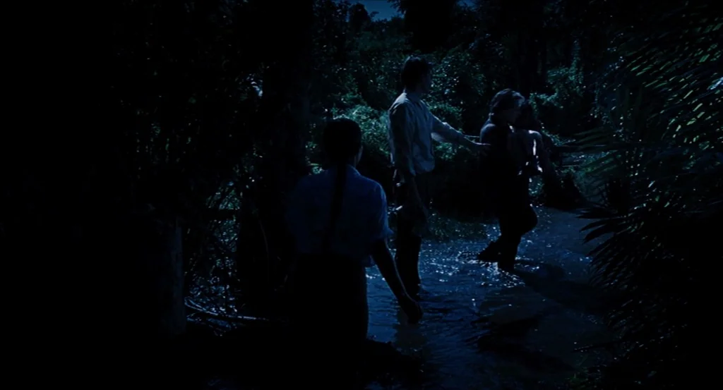 Almayer’s Folly (2011) movie still: fullbody — Three figures wade through a shallow stream in a dense, dark jungle at night. The individuals are…