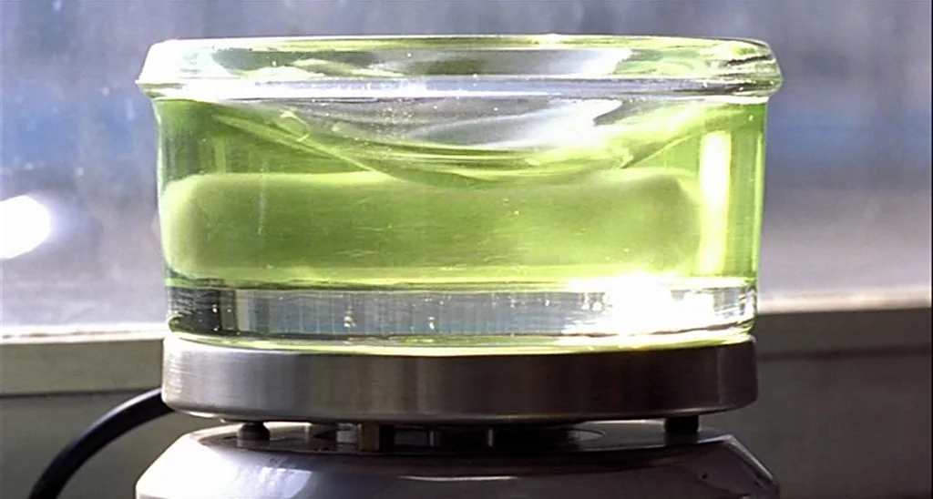 Trouble Every Day (2001) movie still: closeup — A glass container with a pale green liquid rests on a metallic heating plate. A magnetic stir bar…